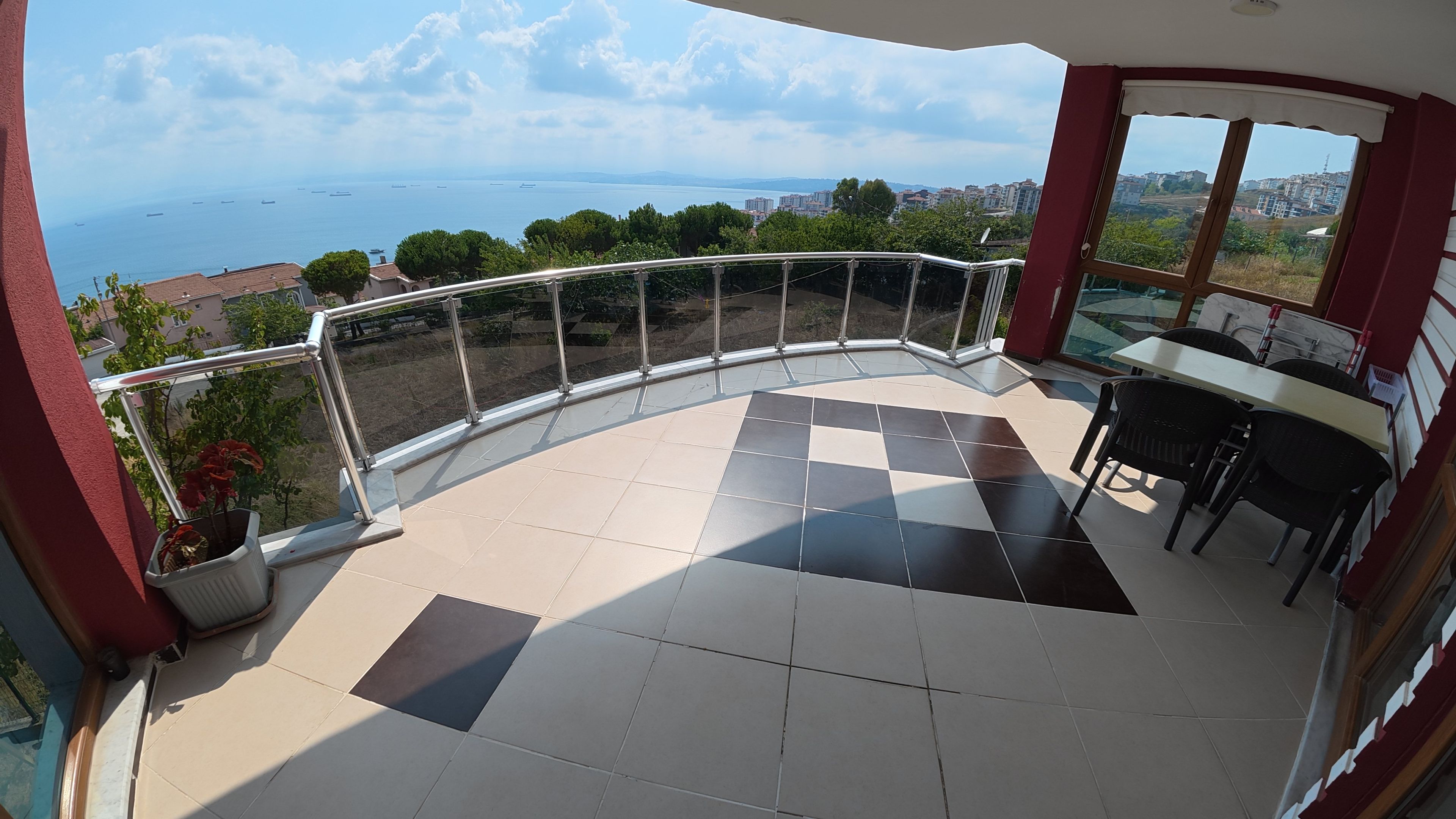 A balcony with a view of the sea and buildings, a table with chairs, and potted plants.