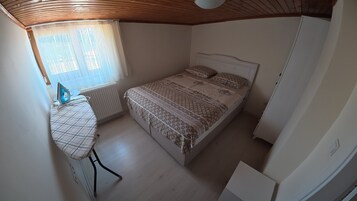 Un dormitorio con cama, tabla de planchar, radiador y ventana con cortinas.