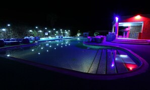 A nighttime swimming pool area with lounge chairs and a small building with a red-lit entrance.