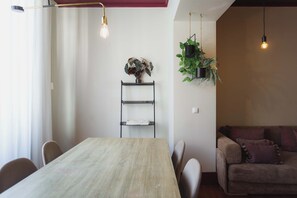 A dining area with a wooden table, chairs, a metal shelf with folded linens, and hanging plants.