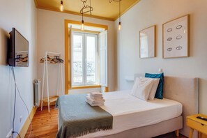 A bedroom with a bed, a bedside table, a TV, a window, and a coat rack.