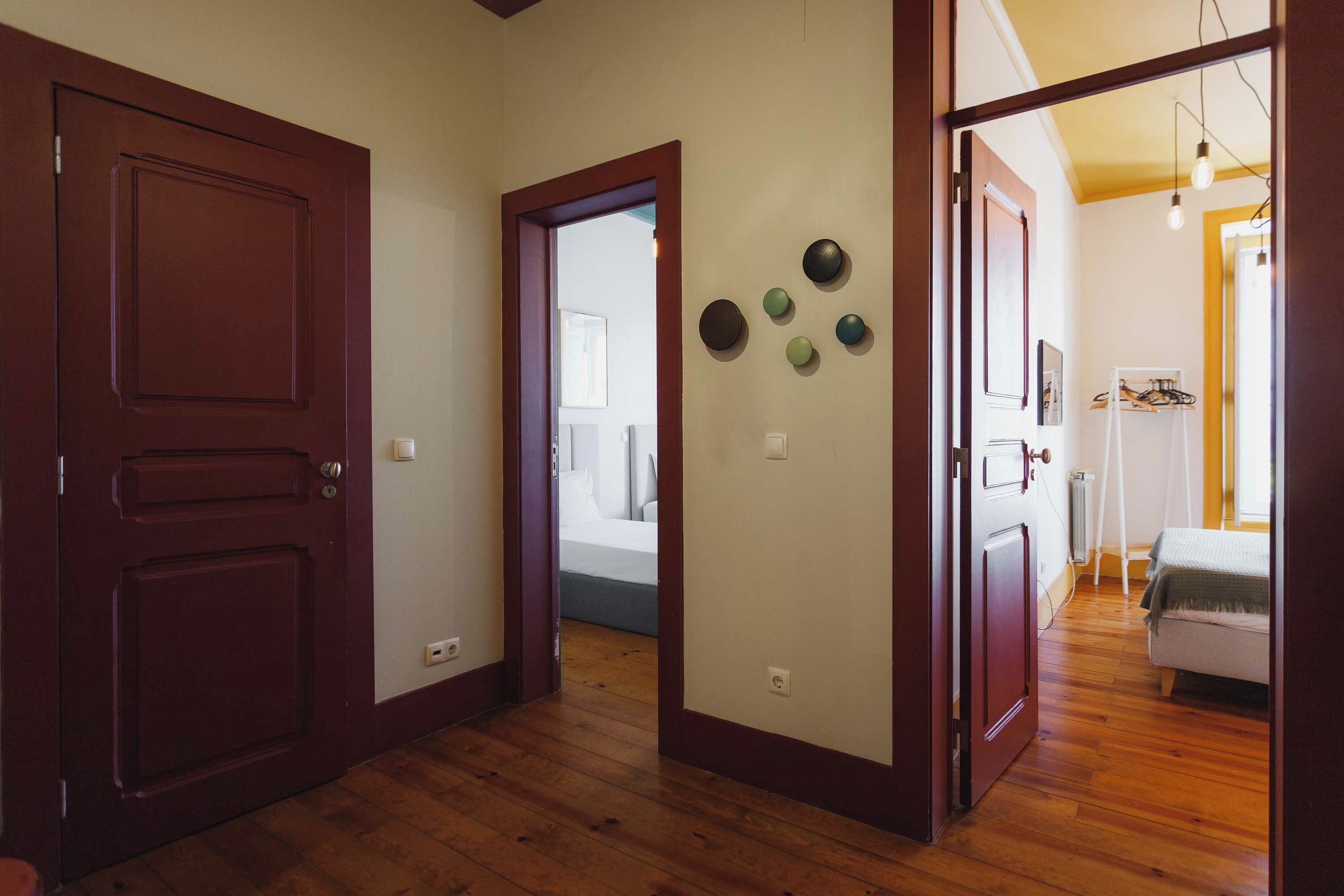 A hallway with wooden flooring, a door on the left, and a bed with a white headboard in the background.