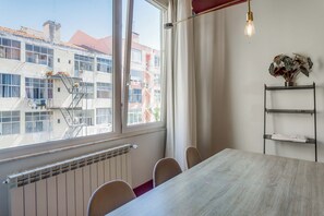 A room with a table and chairs, a radiator, a window with curtains, and a view of buildings outside.