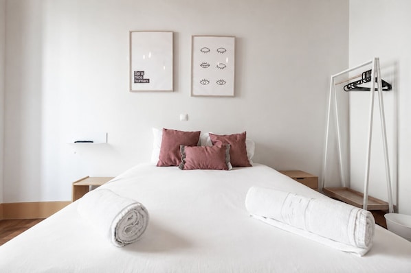 Uma cama bem arrumada com almofadas, uma mesinha de cabeceira, um porta-toalhas fixado na parede e duas fotos emolduradas.