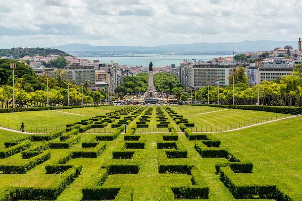 A cityscape with a central monument, surrounded by a well-maintained garden with geometric hedges, modern buildings, and a distant body of water.