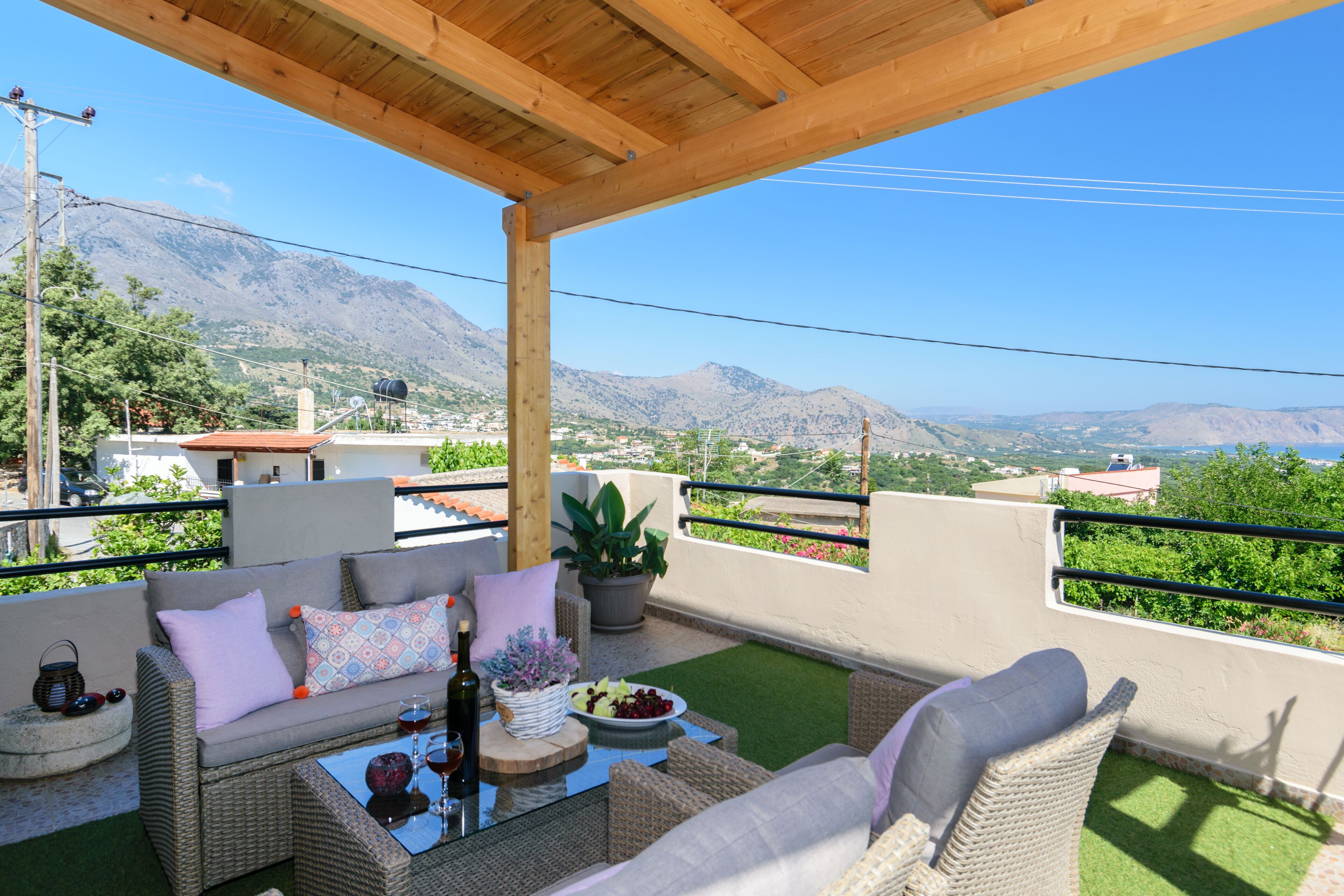 A covered outdoor seating area with wicker furniture, a table with a bottle and glass, and a bowl of fruit.