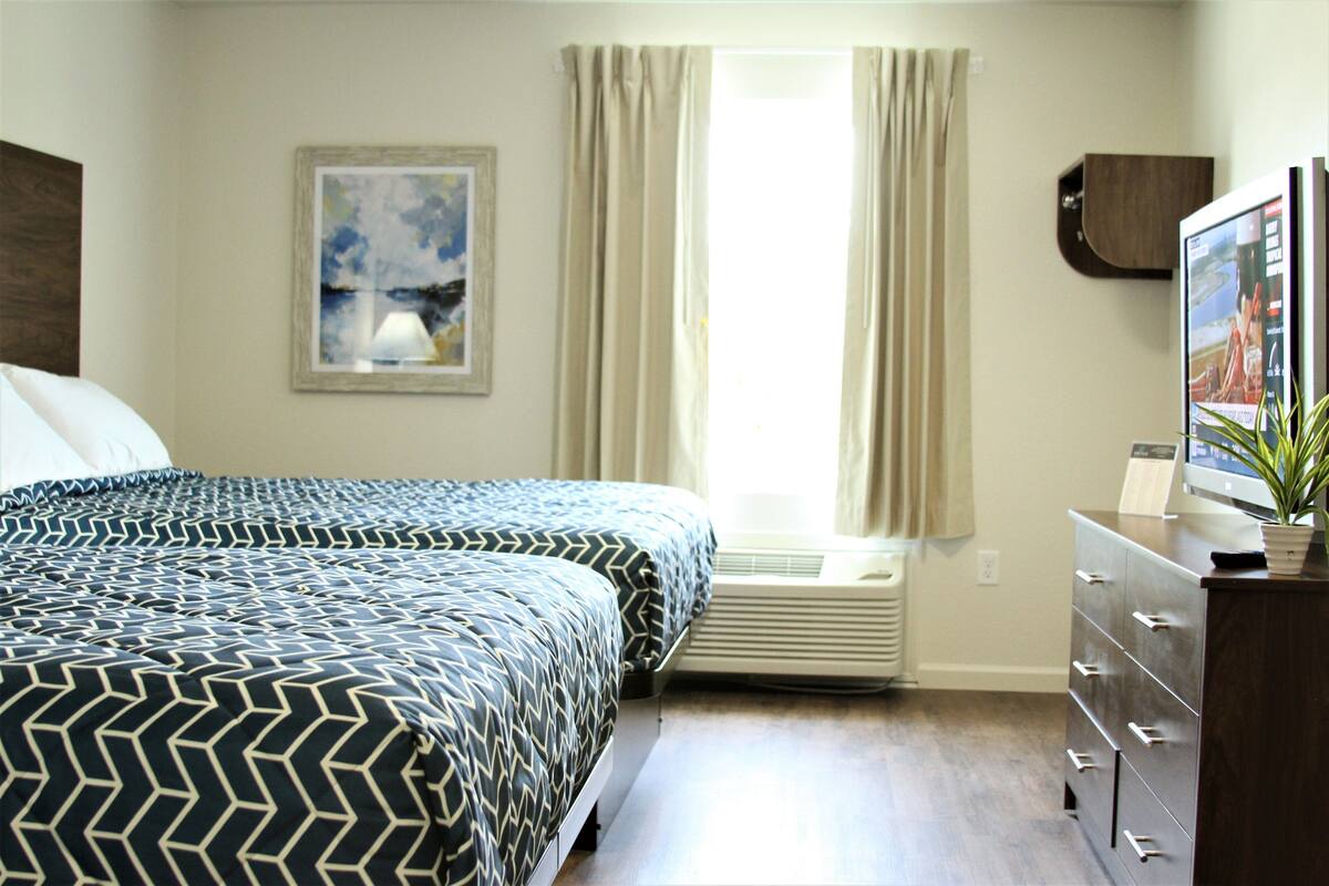 Une chambre à coucher comprenant un lit, un climatiseur, une commode, un téléviseur et une fenêtre avec des rideaux.