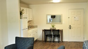 A compact living space with a kitchenette, two chairs at a small table, and a door with a bulletin board. - Stay Plus Extended Stay Suites (Statesboro)