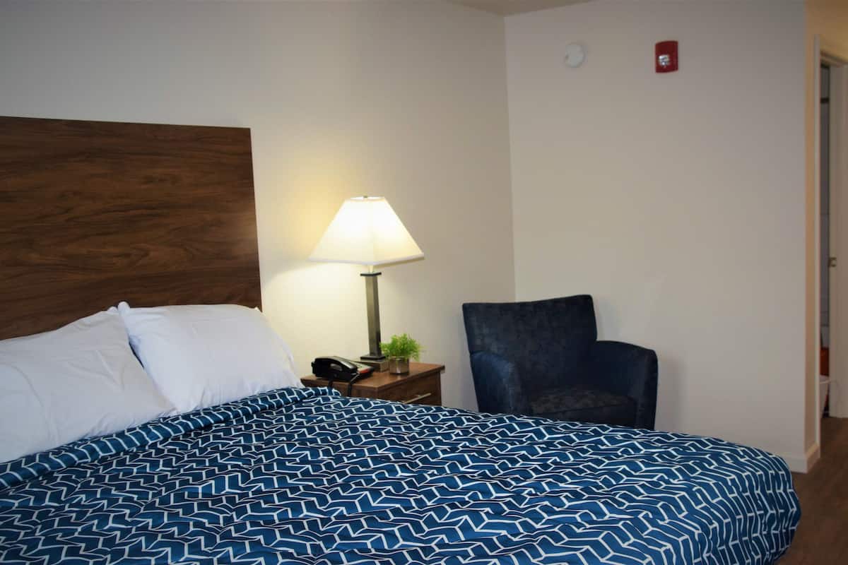 Un lit bien fait, recouvert d’une couvre-lit bleue et blanche à motifs, une lampe de chevet, une chaise et une porte sur laquelle est apposé un panneau rouge indiquant la sortie de secours.