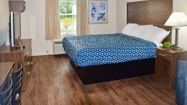A hotel room with a large bed, a desk, a chair, a lamp, and a window with curtains. - Stay Plus Extended Stay Suites (Statesboro)