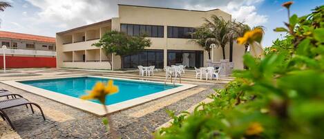 A pool area with a rectangular pool, white lounge chairs, and a building with multiple balconies in the background.