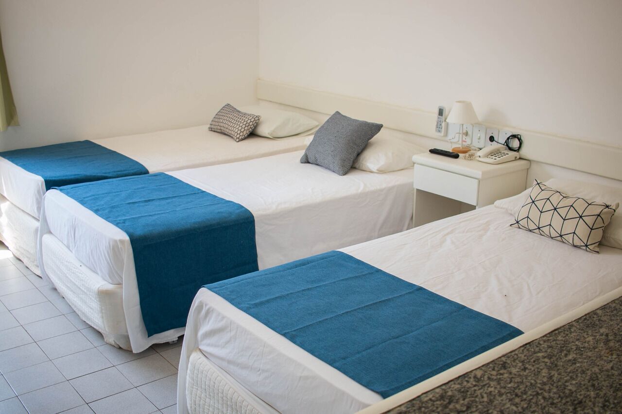 A hotel room with two single beds, each with a blue bedspread and white bedding. There is a bedside table with a lamp, phone, and a small clock. The room has white walls and a tiled floor.