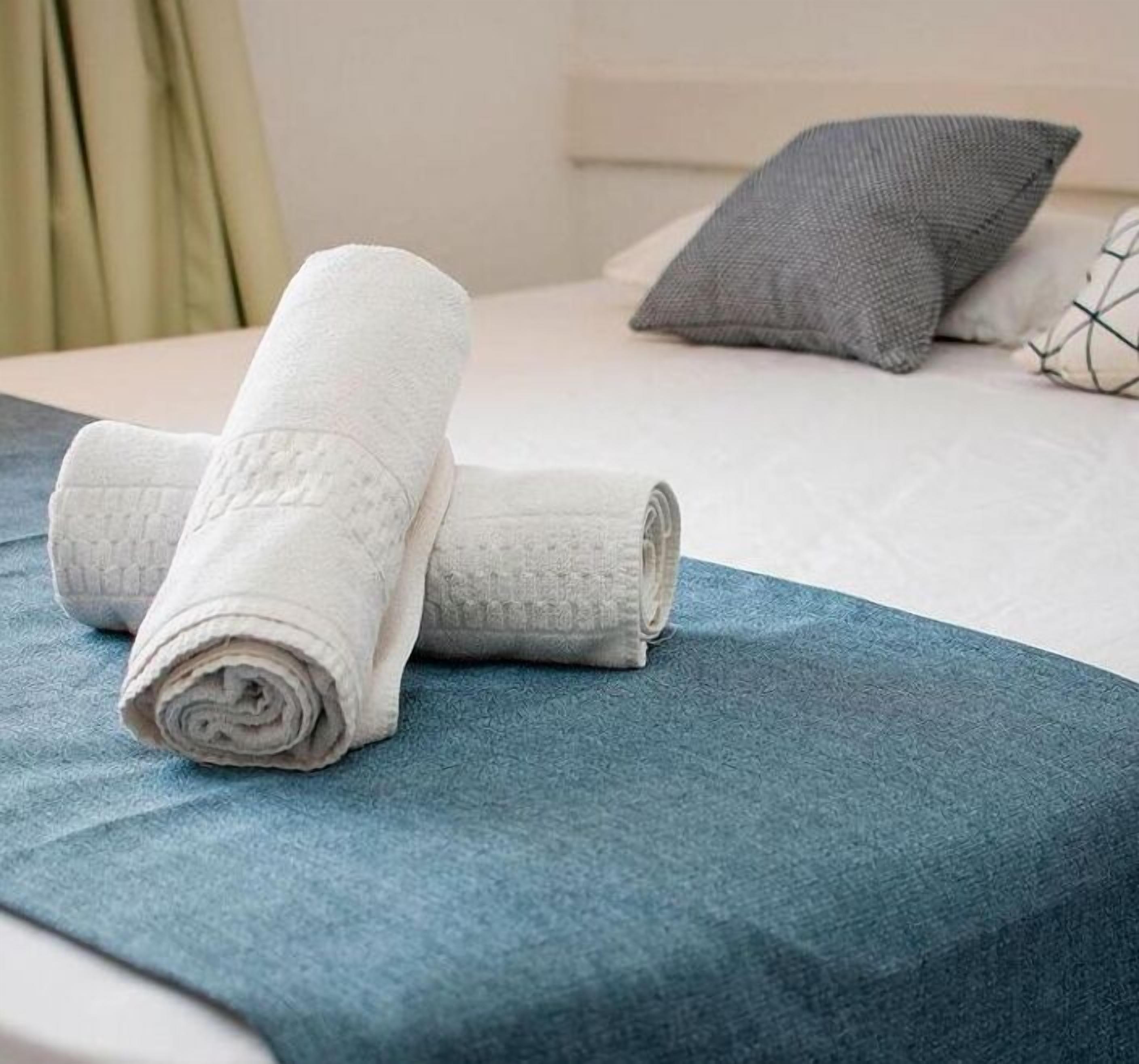 A neatly made bed with a blue bedspread, a white rolled towel, and a grey pillow.