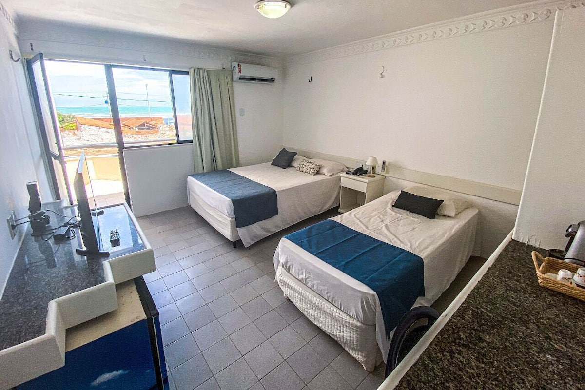 A hotel room with two beds, a desk with a computer, a window with a view, and a small table with a basket.