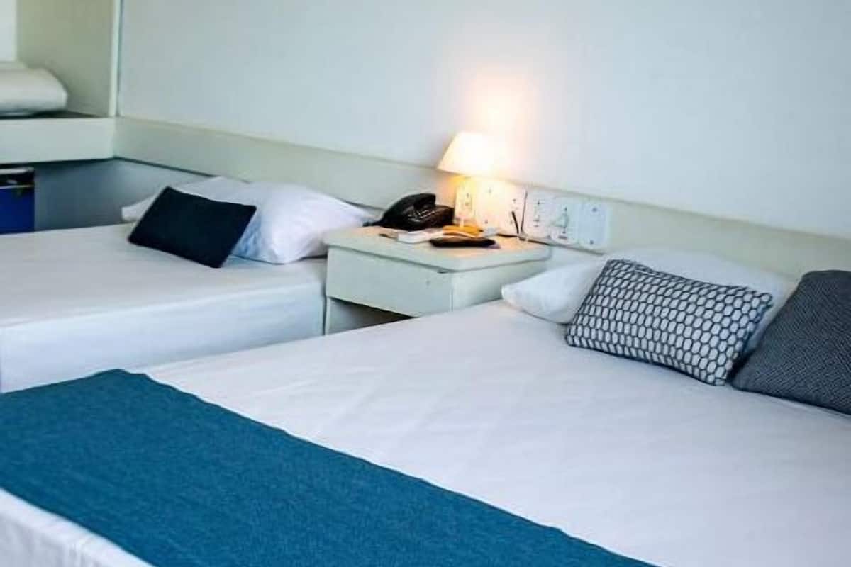 A hotel room with two single beds, each with a white mattress and a blue blanket. There is a nightstand with a lamp, a telephone, and a small table. The room has a simple, clean design with white walls and a tiled floor.