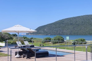 Un espace piscine doté de chaises longues et d’un parasol, offrant une vue sur une étendue d’eau et un paysage vallonné.