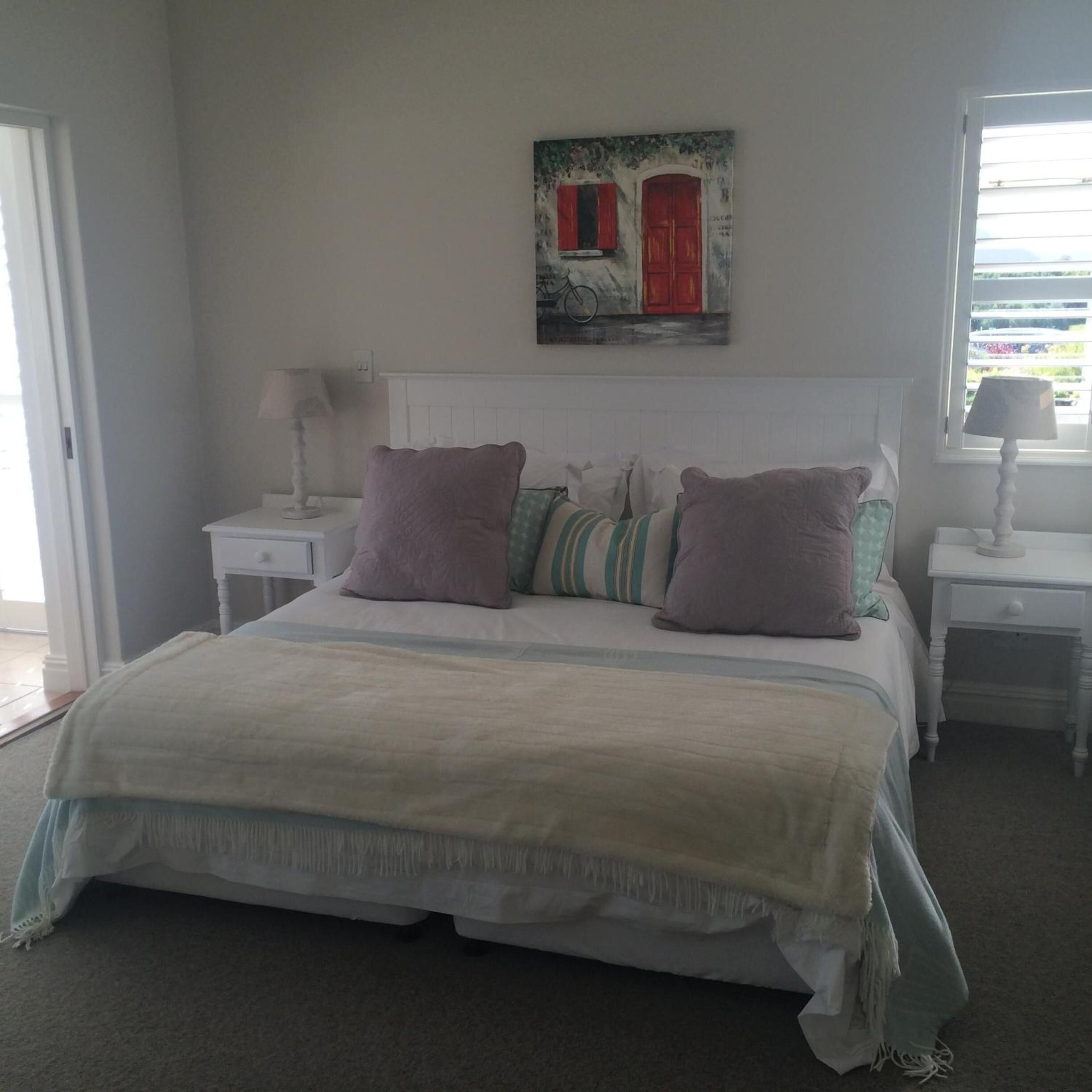 A bedroom with a large bed, white headboard, and two bedside tables with lamps. A painting of a red door and a bicycle is on the wall.