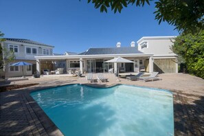 A swimming pool with lounge chairs and umbrellas, surrounded by a paved patio and a house with a covered outdoor area.