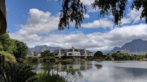 A lakeside hotel with a serene lake, mountains in the background, and lush greenery. - Valley Golf Lodges (Paarl)