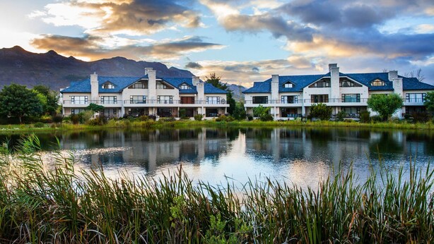 A modern hotel with a large lake in front, surrounded by mountains and lush greenery. - Valley Golf Lodges (Paarl)