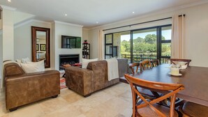 A spacious living area with a fireplace, comfortable seating, a dining table, and large windows offering a view of the outdoors. - Valley Golf Lodges (Paarl)