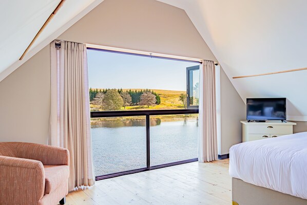 Un dormitorio con un amplio ventanal corredero que da a una vista de un lago, una cama, un televisor sobre una mesita de noche y una silla.