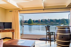 A room with a TV, a wooden desk, a chair, a wooden barrel, and a view of a lake and trees.