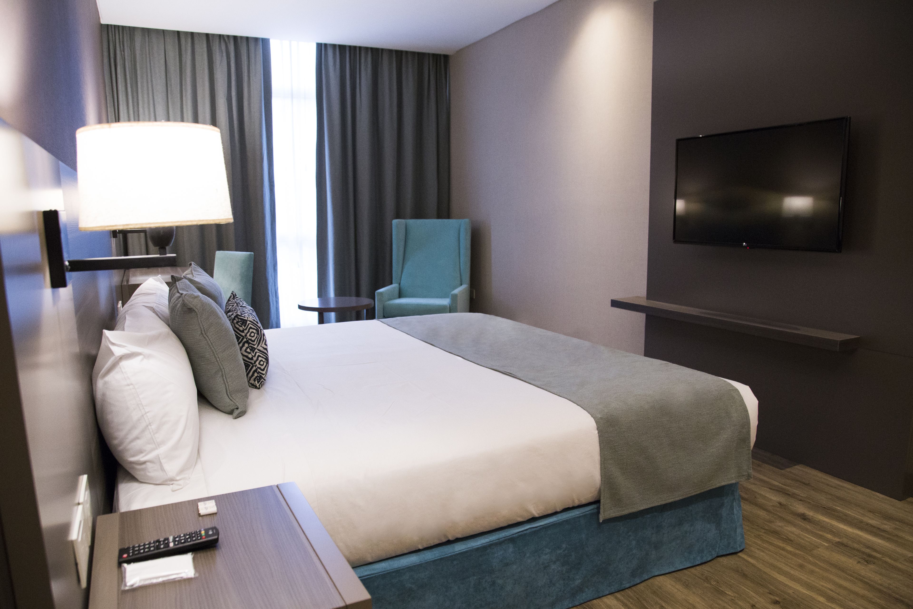 A hotel room with a bed, a desk with a lamp, a chair, a TV mounted on the wall, and a window with curtains.