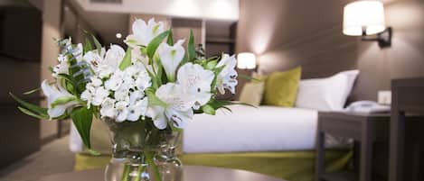 Una habitación de hotel con cama, mesita de noche, lámpara y un jarrón con flores blancas sobre una mesa redonda.