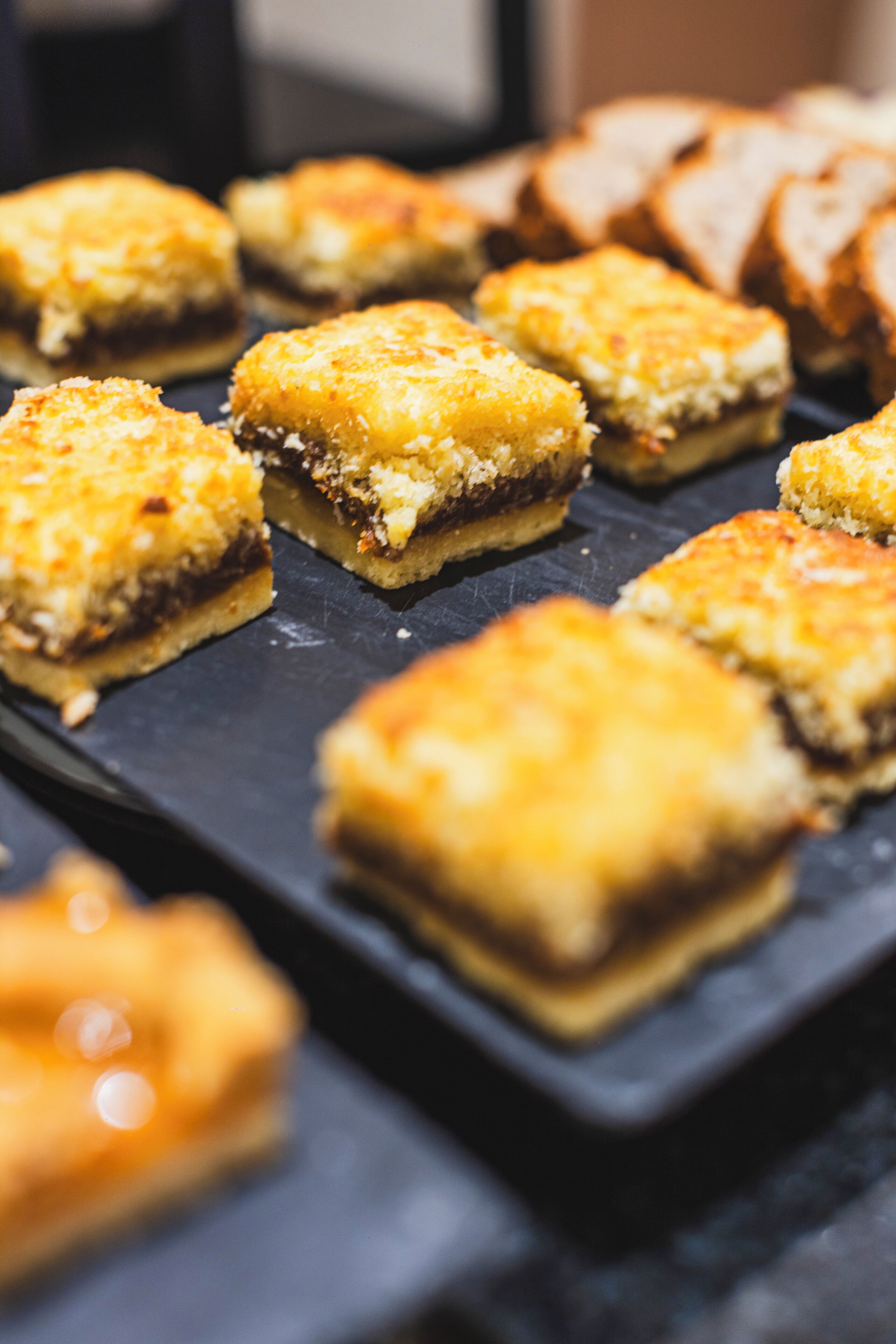 Une assiette contenant des desserts carrés à la croûte dorée, peut-être un type de gâteau ou de pâtisserie, posée sur une surface sombre en ardoise.