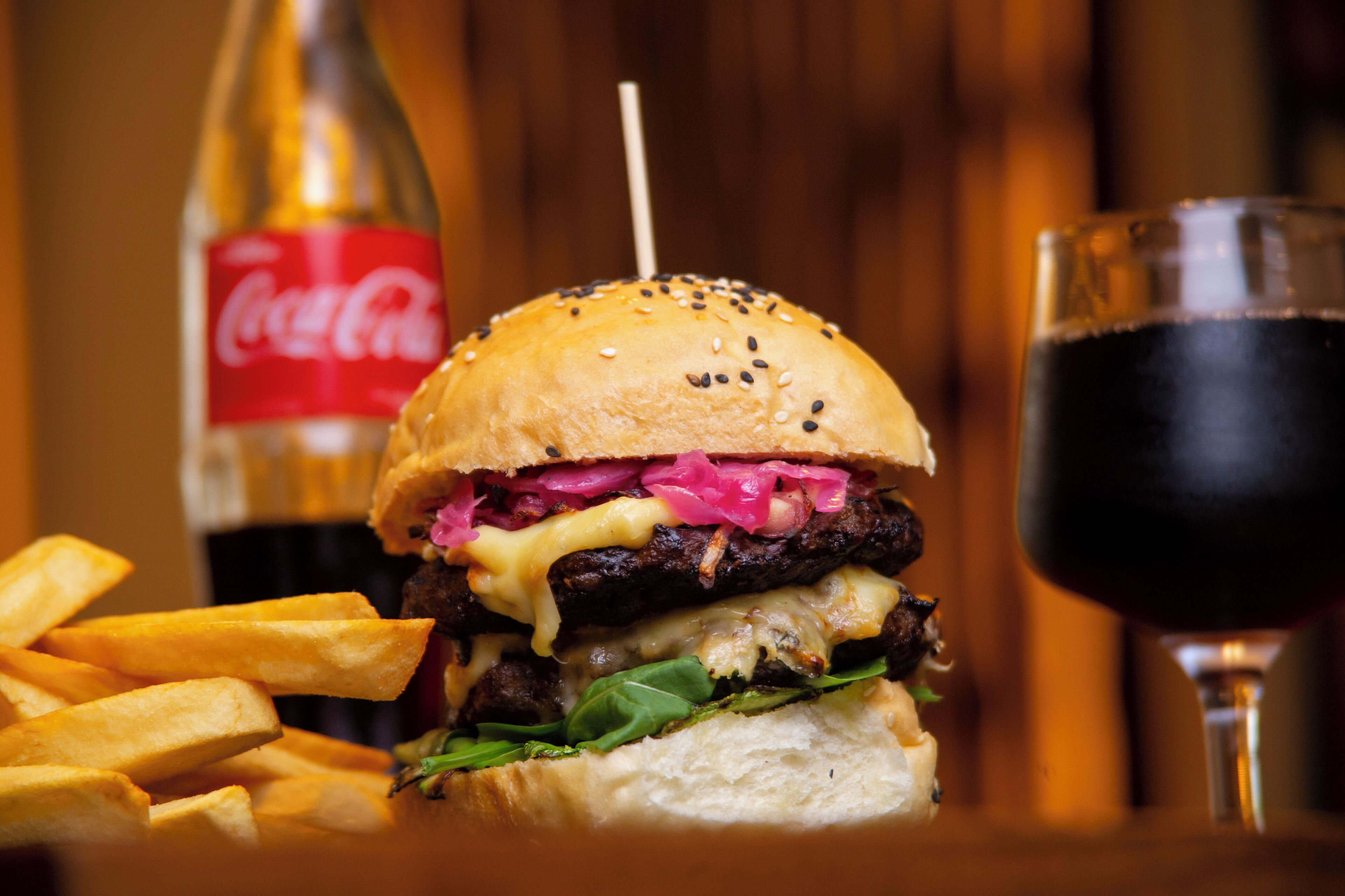 Una hamburguesa con papas fritas y un vaso de bebida oscura, posiblemente Coca-Cola, sobre una mesa de madera.