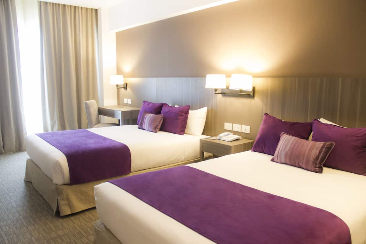 Een hotelkamer met twee bedden, elk met een paarse sprei, een nachtkastje met een lamp en een bureau met een stoel.