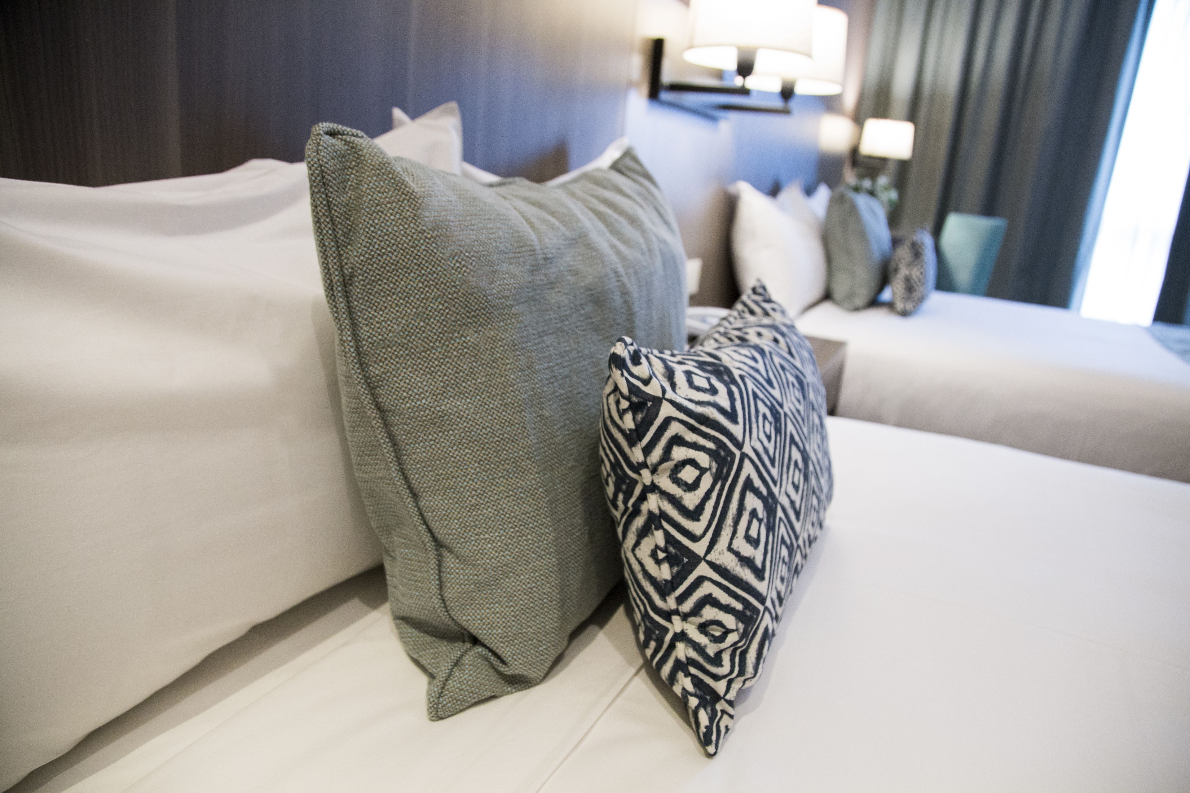A neatly arranged bed with a grey and a patterned pillow, a bedside table with a lamp, and a view of a window with curtains.
