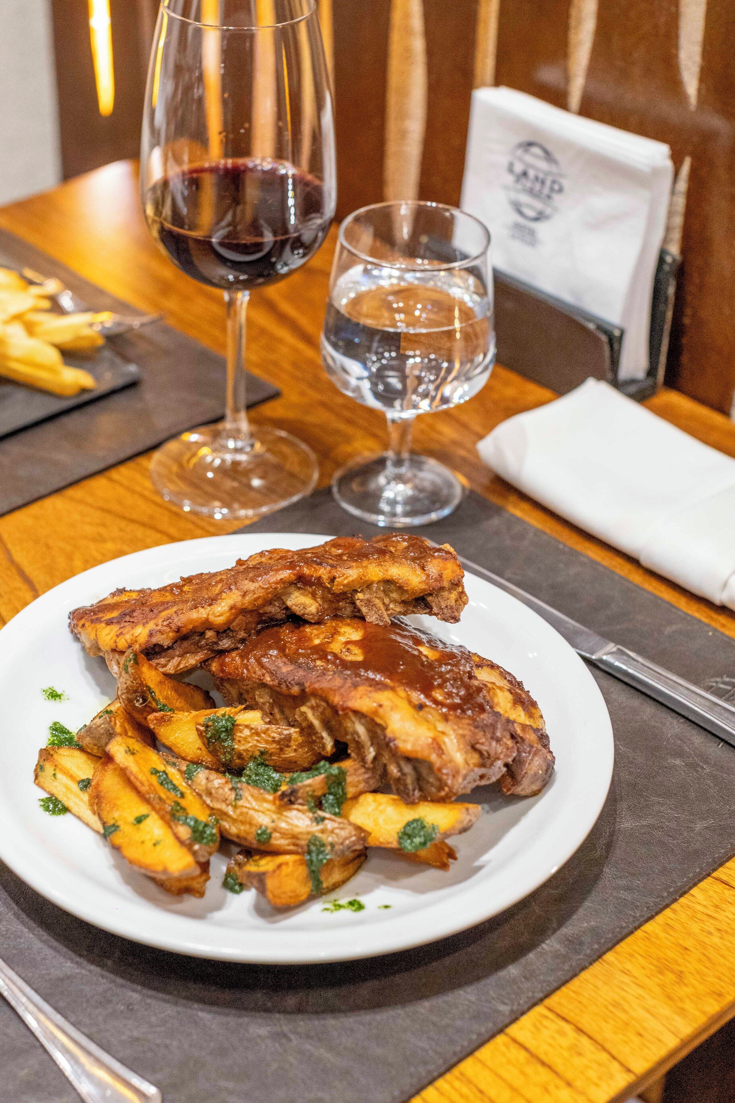 Un plato de costillas y papas fritas con una copa de vino tinto sobre una mesa de madera.