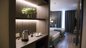 Ein Hotelzimmer mit einem hölzernen Schreibtisch, einem Wasserkocher, Tassen und einem Blumenstrauß. Ein Bett mit blauer Bettdecke und ein Fenster mit Vorhängen sind ebenfalls vorhanden.