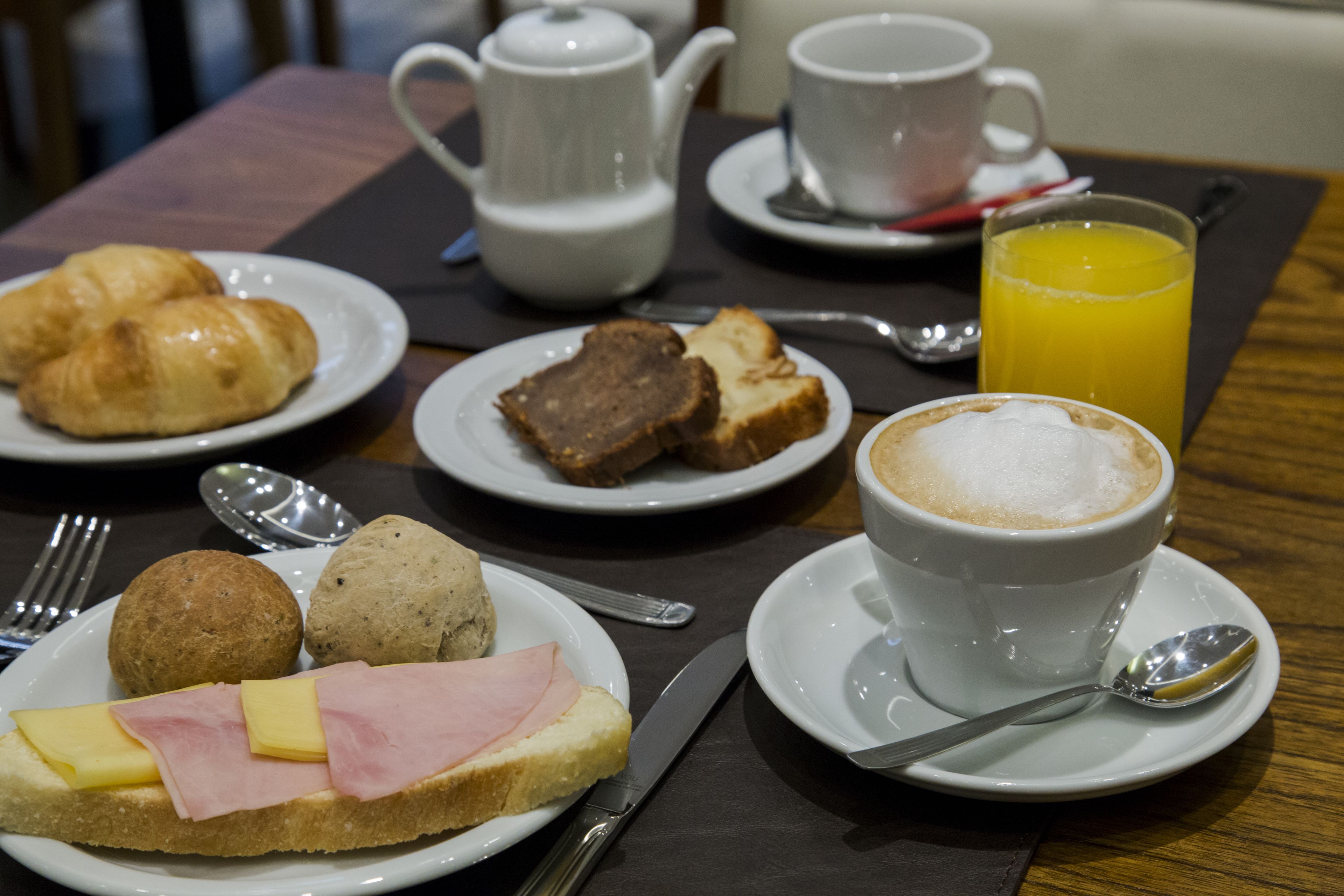 A breakfast setting with croissants, ham, cheese, muffins, and a cup of coffee with a glass of orange juice on a table.