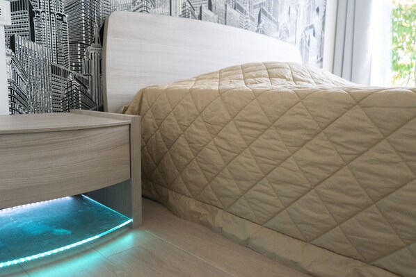 Una cama con cubrecolcha beige acolchado, una mesita de noche de madera con luz LED integrada y un papel tapiz con motivos de paisaje urbano.