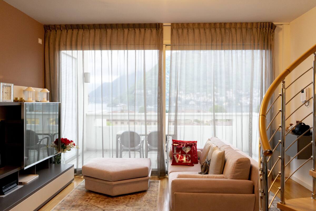 A modern living room with a sofa, ottoman, and TV stand. There is a large window with sheer curtains, a view of the city, and a staircase.