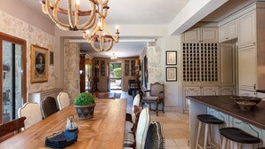 Un vaste espace dédié à la salle à manger, avec une table en bois, des chaises et un lustre. On y trouve également un meuble à vin et un coin cuisine équipé de tabourets de bar.