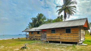 A wooden cabin with a metal roof, situated on a grassy area near a body of water, with palm trees in the background. - Private wood Cabins on San Blas island - private bathroom (Narganá)