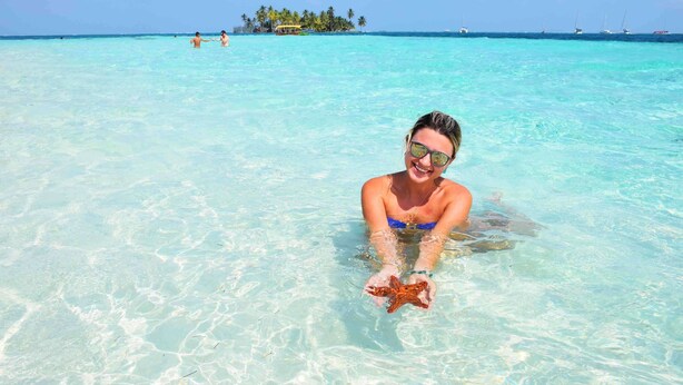 A person holding a starfish in clear turquoise water with a distant island and boats in the background. - Private wood Cabins on San Blas island - private bathroom (Narganá)