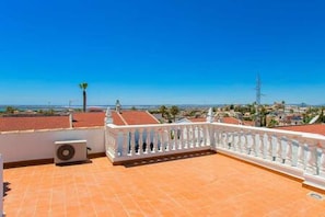 Um terraço no telhado com balaustrada branca, piso de telhas vermelhas e vista clara da paisagem urbana e do litoral.