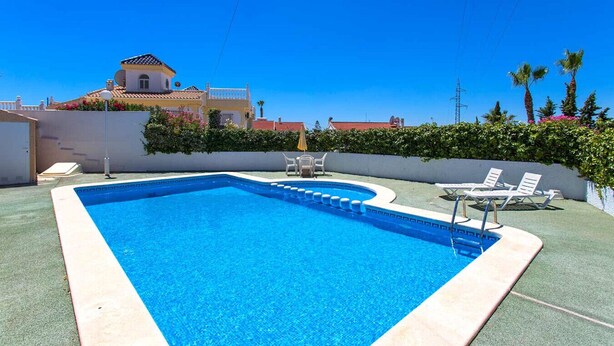 A rectangular swimming pool with a tiled edge, surrounded by a green deck. There are lounge chairs and a small table near the pool.
