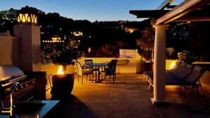 Eine Terrasse mit Grill, Stühlen und Tisch, mit Blick auf eine Stadtlandschaft im Abenddämmerung.