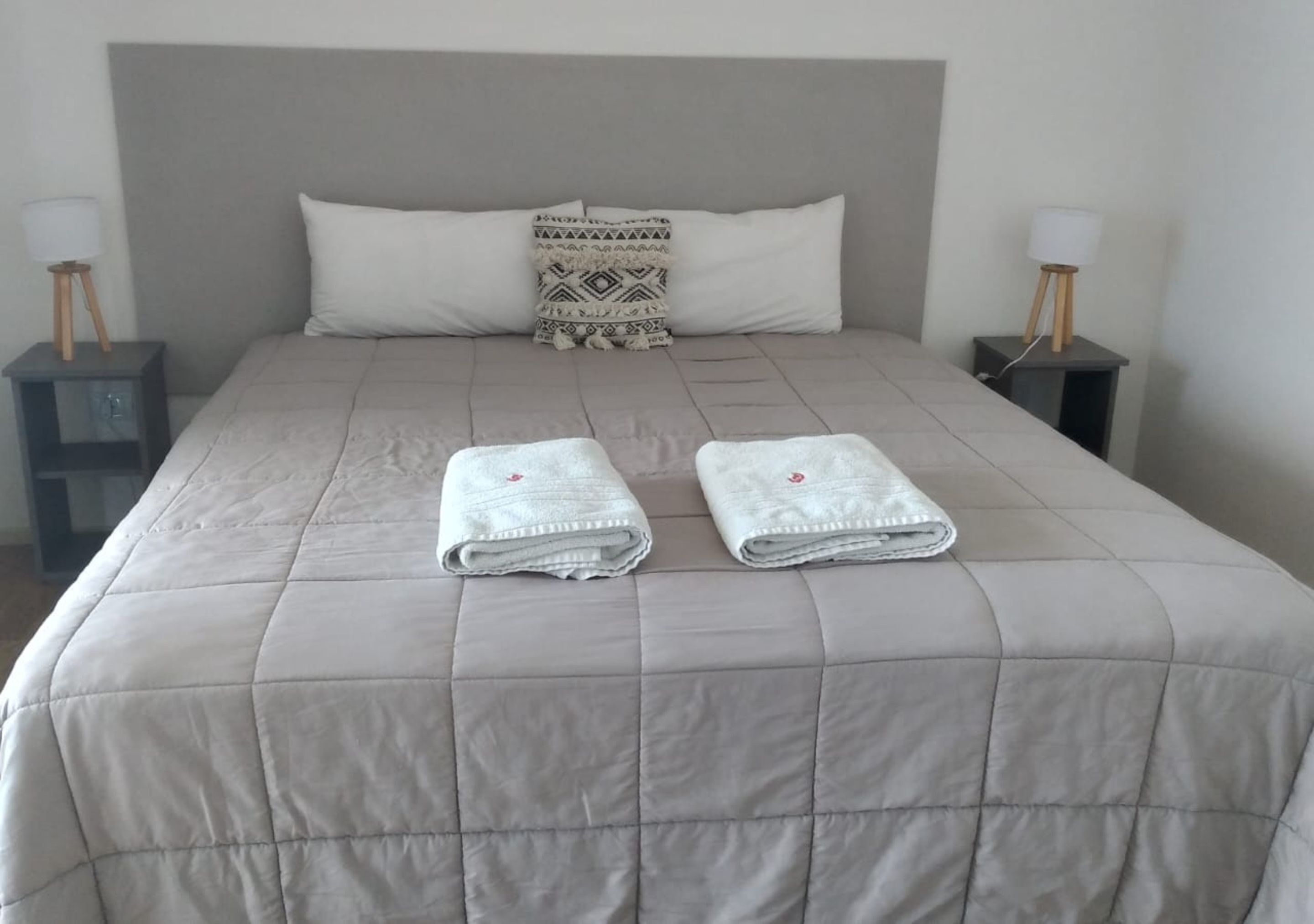 A neatly made bed with a quilted grey duvet, two folded white towels, a patterned throw pillow, and two bedside tables with lamps.