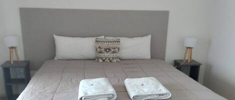 A neatly made bed with a quilted grey duvet, two folded white towels, a patterned throw pillow, and two bedside tables with lamps.