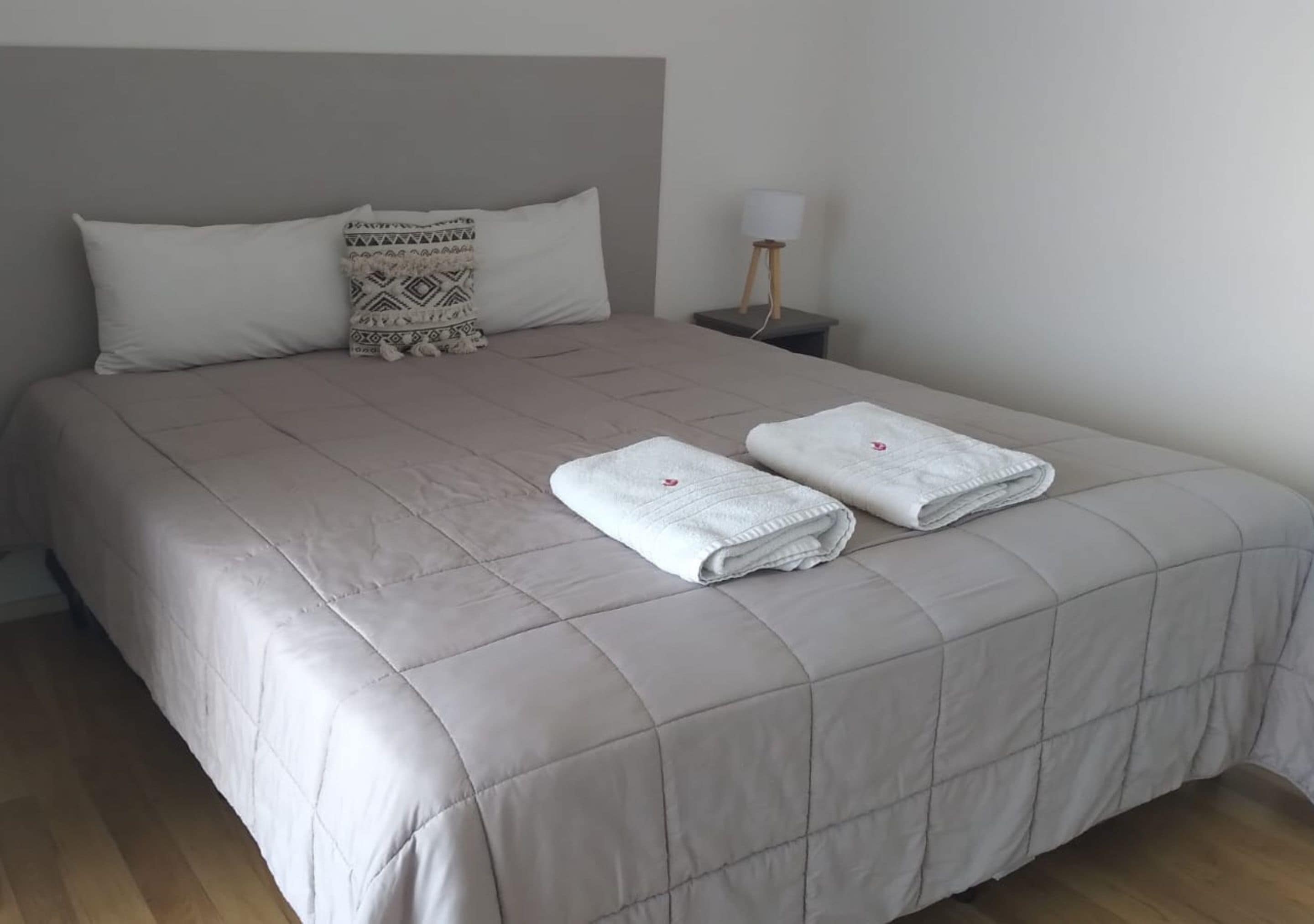 A neatly made bed with a grey quilt, two folded white towels, and a patterned pillow.