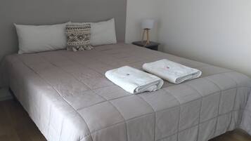 A neatly made bed with a grey quilt, two folded white towels, and a patterned pillow.