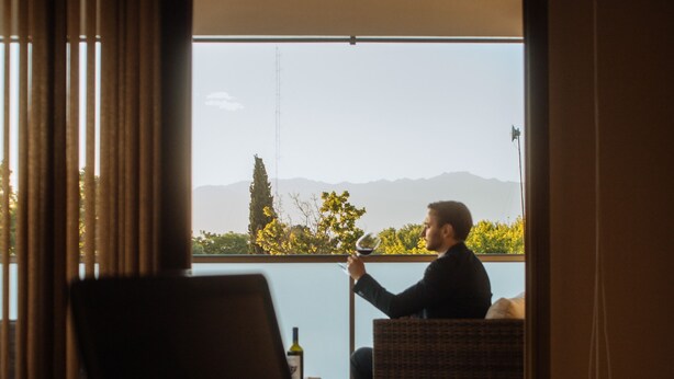 Une chambre d’hôtel avec un lit, un ordinateur portable posé sur le lit, et une personne tenant un verre de vin sur un balcon d’où l’on peut admirer des montagnes et des arbres.