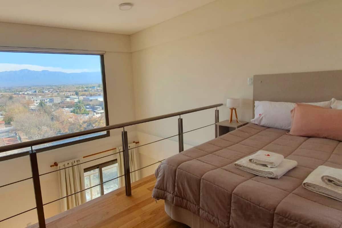 Une chambre avec une grande fenêtre, un lit recouvert d’une couette marron, et une vue sur le paysage urbain.
